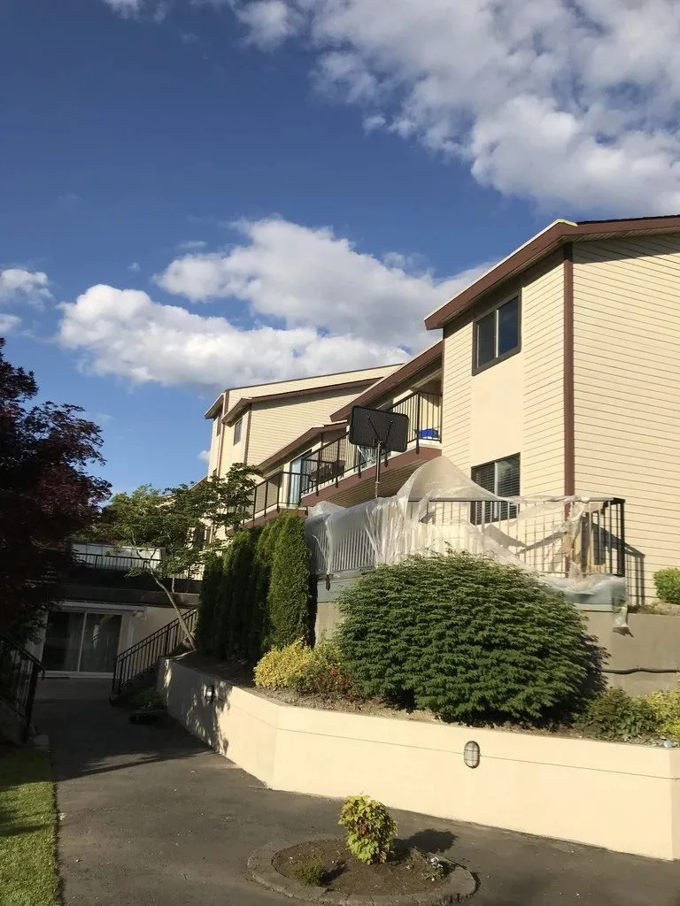 A townhouse complex in Burnaby photographed mid-job: balconies and railings wrapped, planting covered, the elevation ready for coating. People live here while the work goes on, so the masking is part of the job rather than an afterthought.