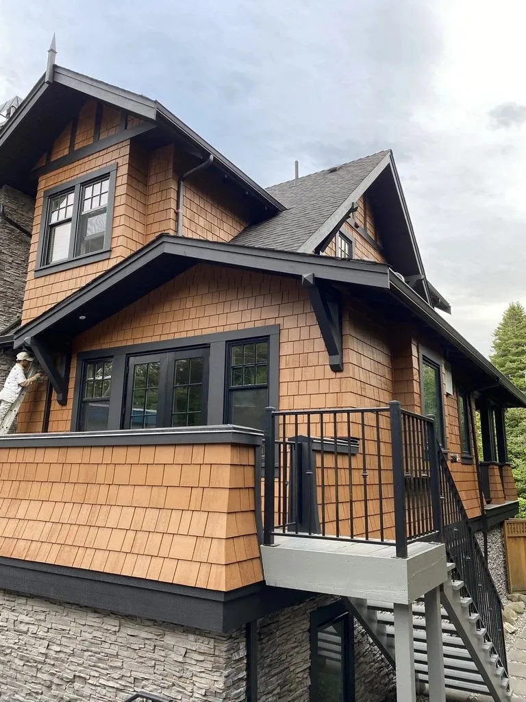 A North Vancouver house in cedar shingles, kept in a warm natural tone, with fascia, brackets, window frames and deck rails carried in charcoal. A Mayda painter is at work on the left of the frame. Two finishes on one house, and they do not age at the same rate.