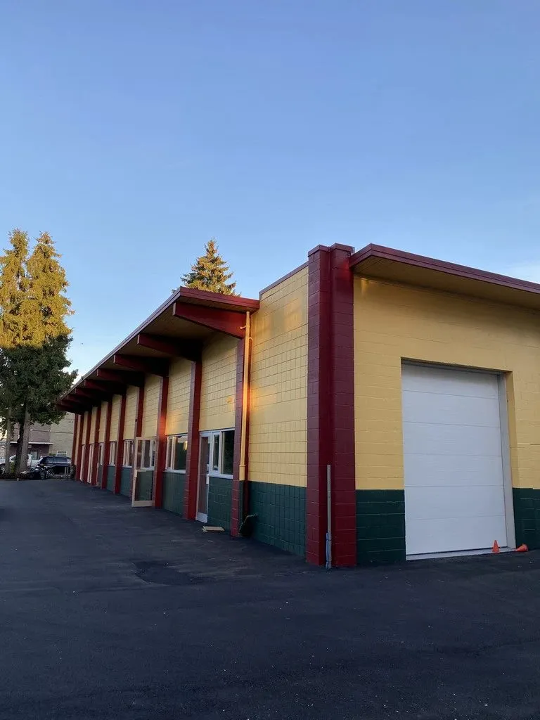 A commercial building in Burnaby, in painted concrete block: a yellow body, deep red columns and fascia, a dark green base course, and a white roll-up door. Three colours and four materials on one frontage, each needing its own preparation.
