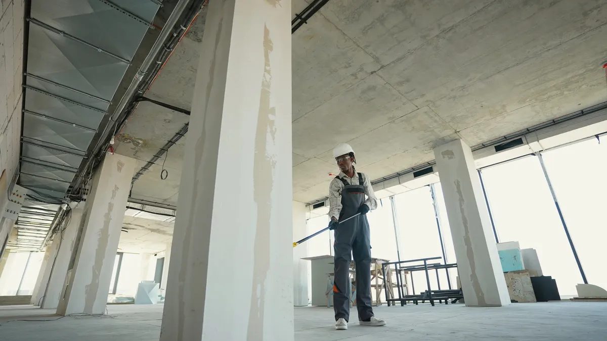 Painter on an empty office floor rolling a ceiling with an extension pole beside tall windows